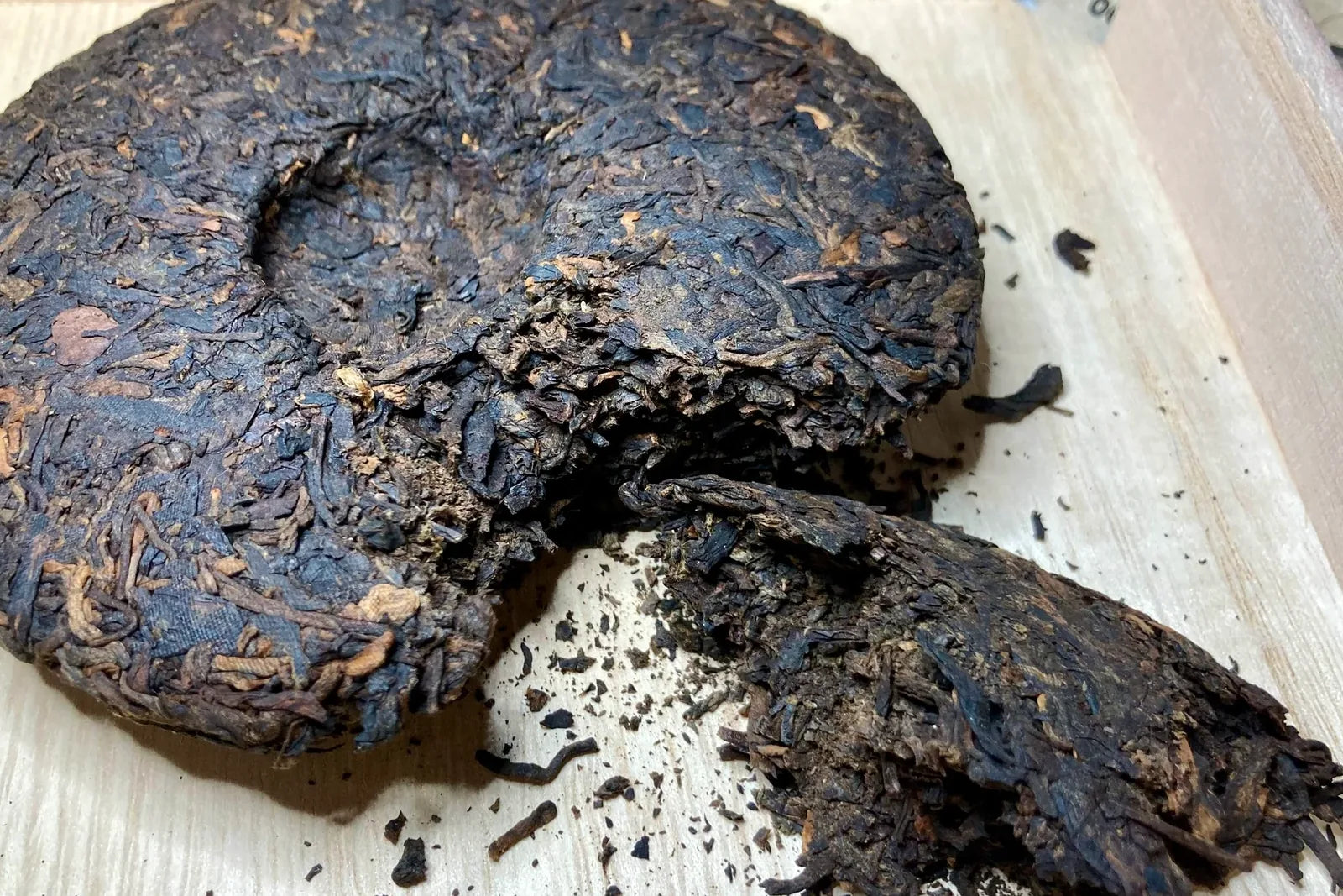 Round dark brown-black aged shu pu-erh tea cake with a chunk broken off on a pale wooden surface