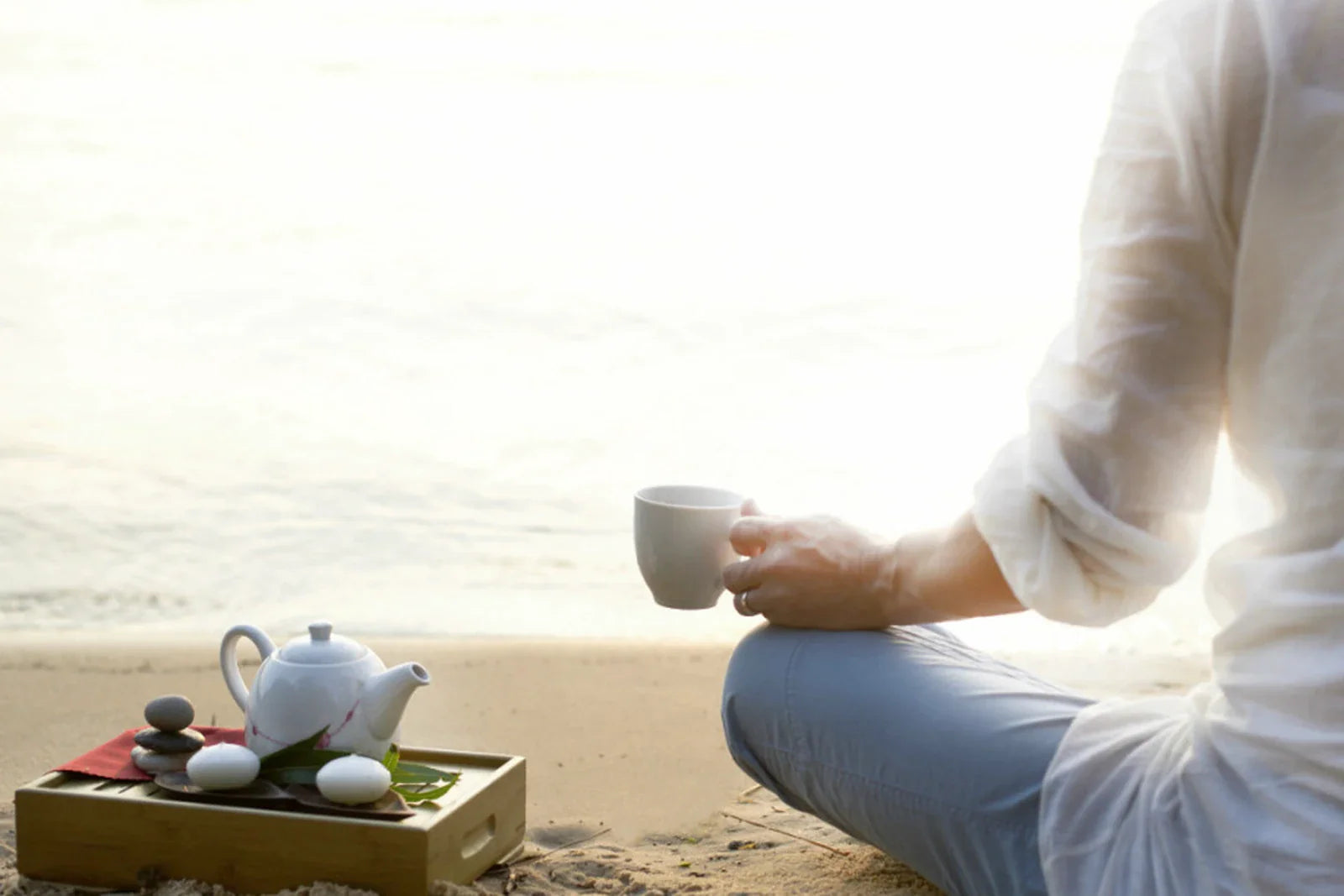 Woman in white sitting cross-legged on a beach at sunrise holding a white tea cup with a small teapot and stacked stones beside her