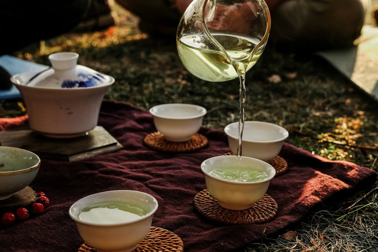 Glass pitcher pouring pale green tea into small white gongfu cups arranged on woven coasters at dusk outdoors