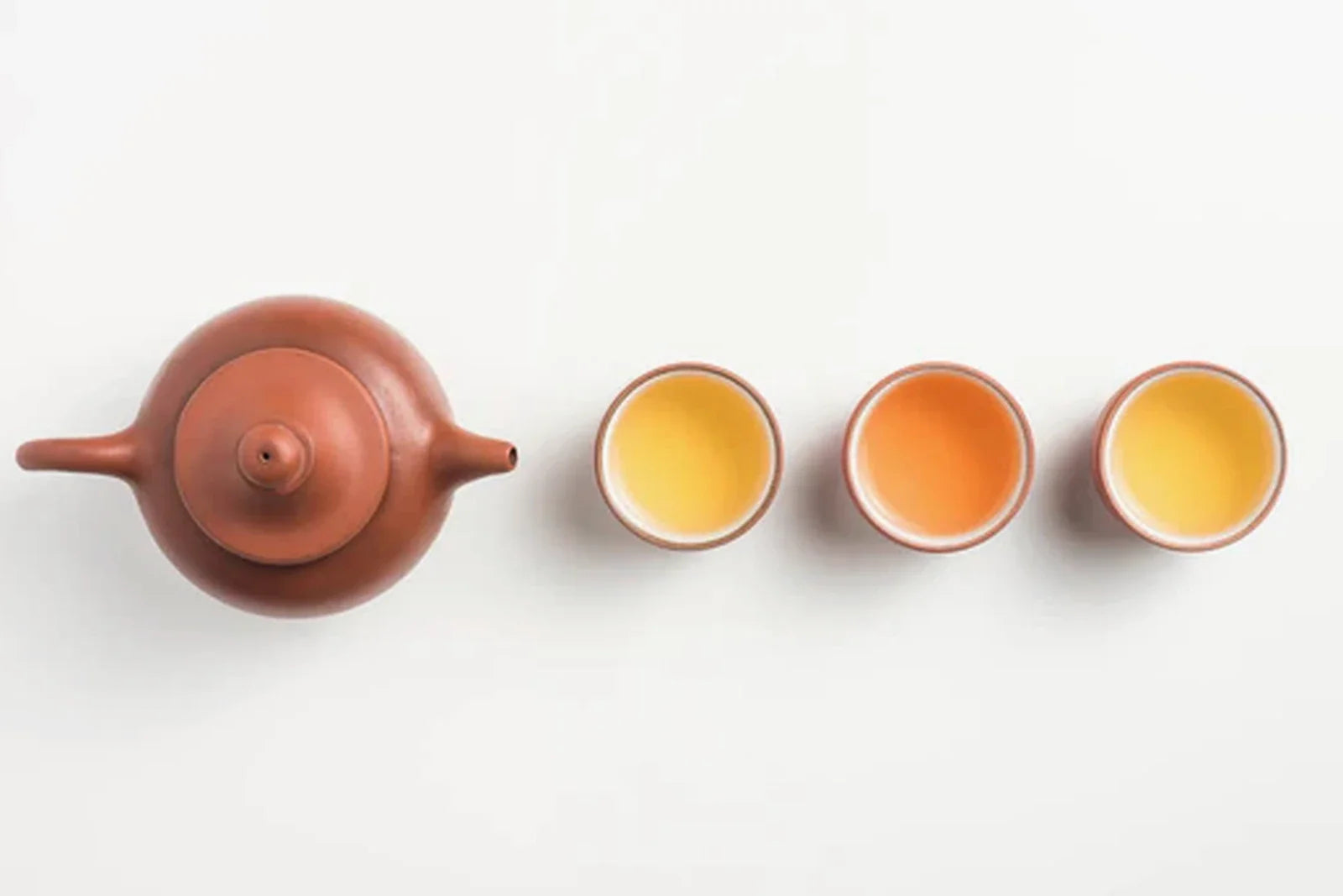 Overhead flatlay of a small Yixing clay teapot beside three small cups of amber oolong tea liquor