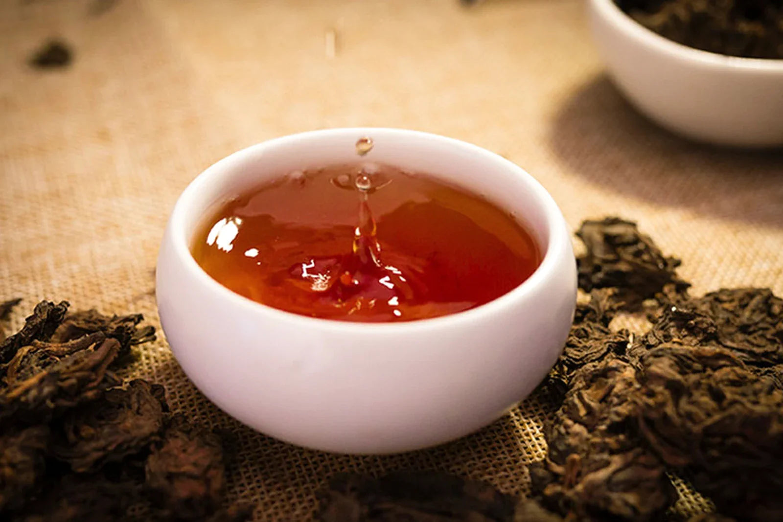 Beautiful close-up of dark red pu-erh tea in a white cup with a water droplet splash surrounded by dry fermented leaves