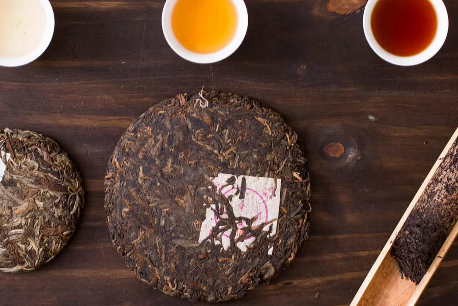 Three pu-erh tea cups showing dark red ripe shu, amber young sheng and pale fresh sheng beside three pressed cakes