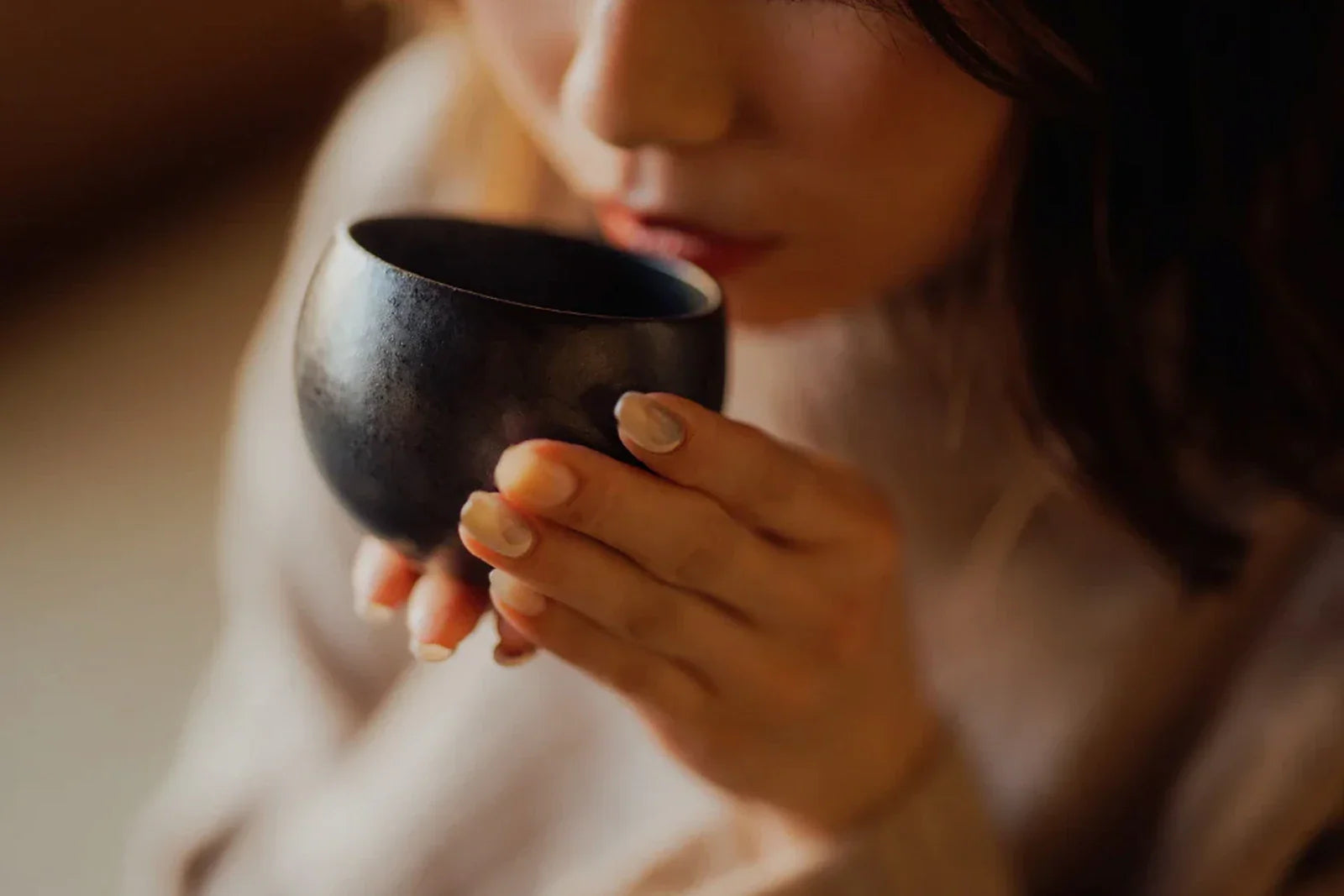 Close-up of a woman in a cream knit sweater sipping evening tea from a small dark ceramic cup in soft warm light