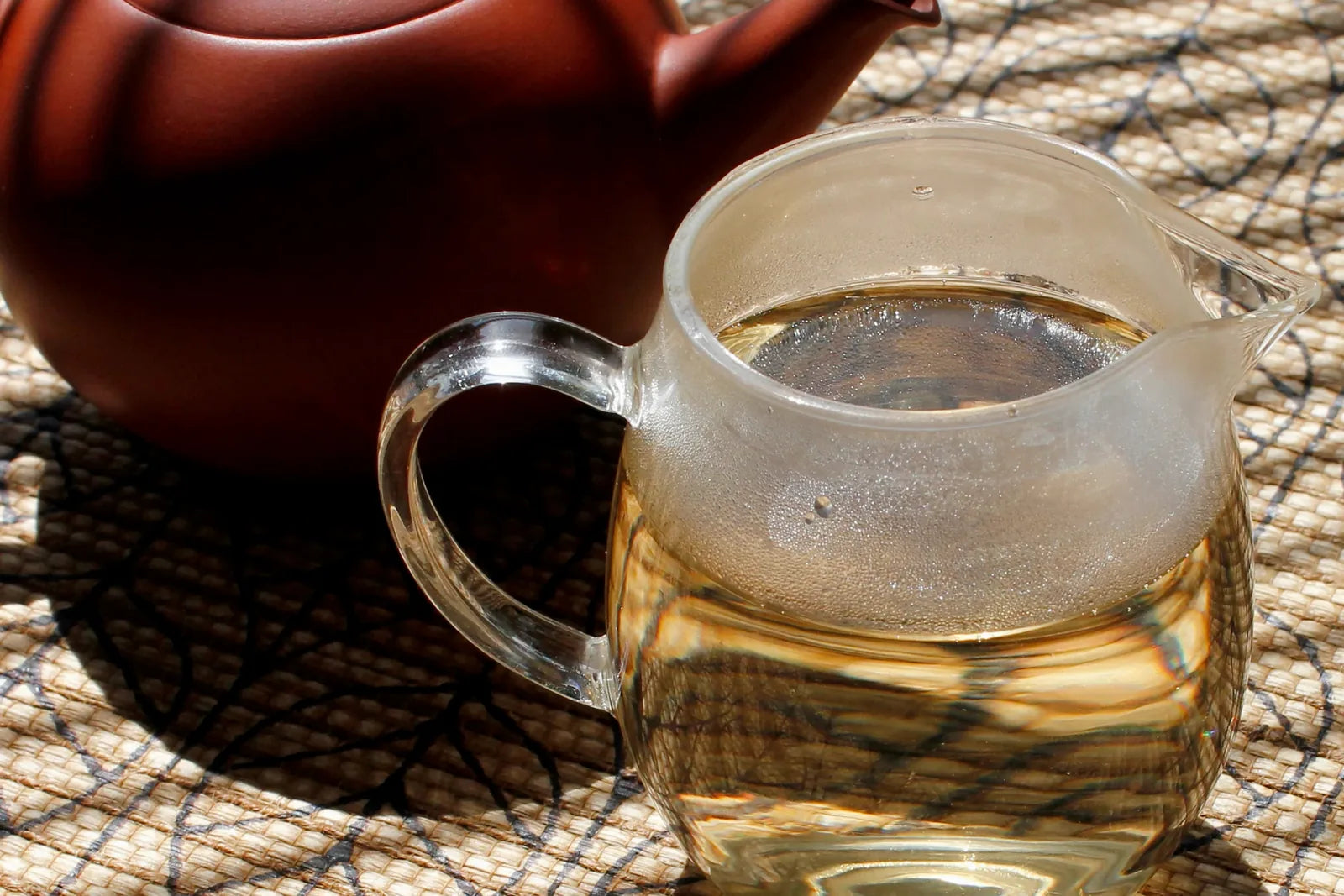 Red clay teapot beside a glass pitcher of pale yellow Moonlight White tea on a sunlit bamboo mat