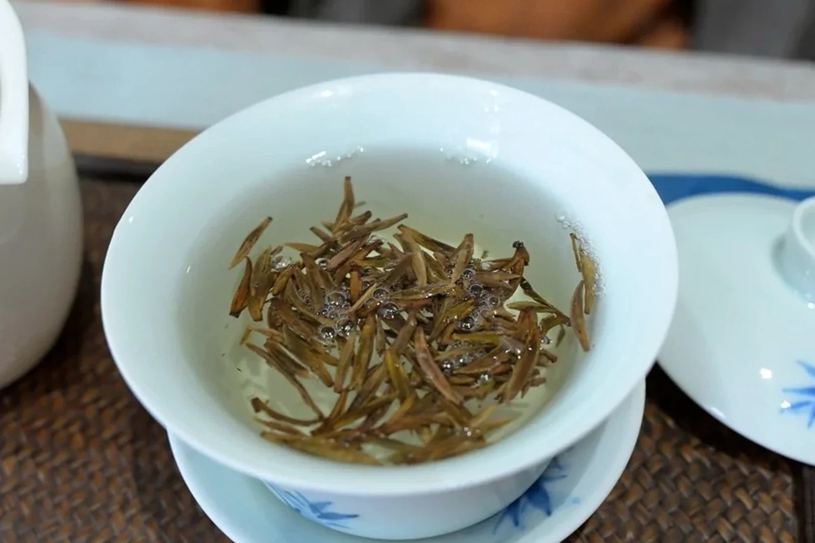 White gaiwan with slender golden-downy yellow tea buds suspended in clear pale liquor on a bamboo mat